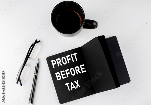 PROFIT BEFORE TAX written text in small black notebook with coffee , pen and glasess on a white background. Black-white style