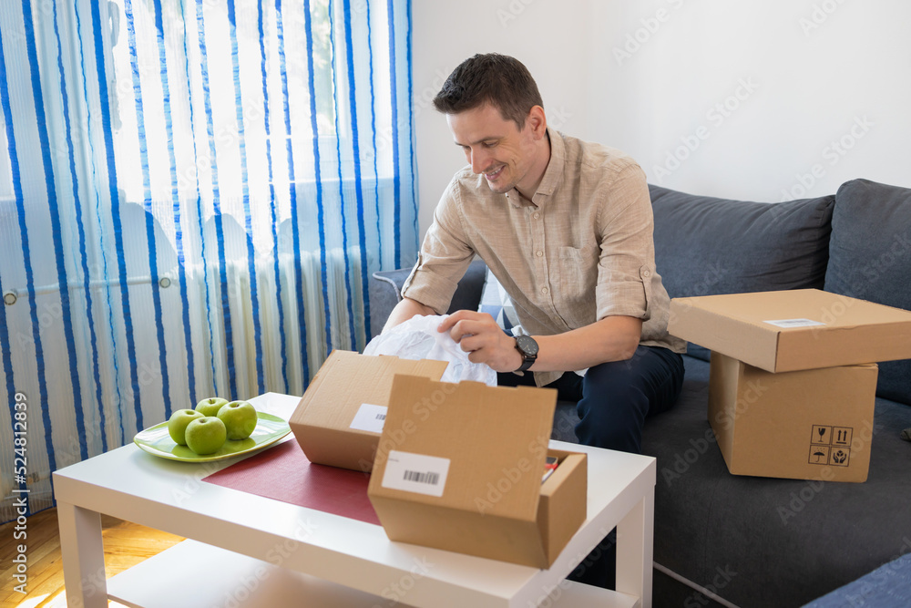 Man unpacking awaited parcel, looking inside, sitting at sofa, satisfied happy customer opening cardboard box with online store order, good shipping delivery service. Man unpacking awaited parcel, looking inside, sitting at sofa, satisfied happy customer opening cardboard box with online store order, good shipping delivery service.