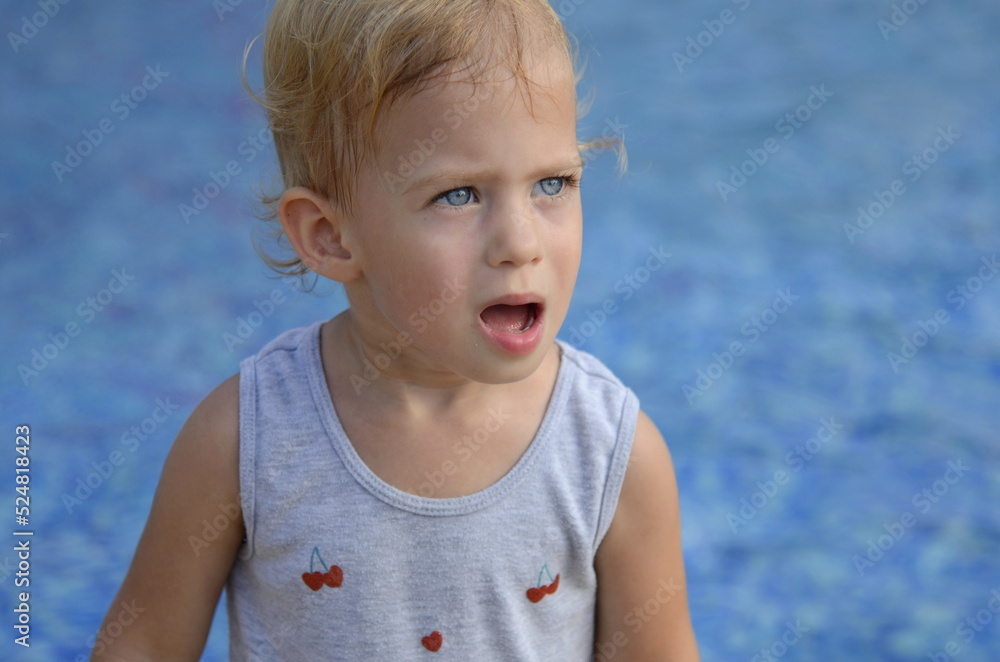 Sweet little girl, two years old. The girl smiles, looks into the camera. Blue eyes, blonde hair. In the background there is a swimming pool, a fountain, blue water. Concept: summer, holidays, traveli Sweet little girl, two years old. The girl smiles, looks into the camera. Blue eyes, blonde hair. In the background there is a swimming pool, a fountain, blue water. Concept: summer, holidays, traveli