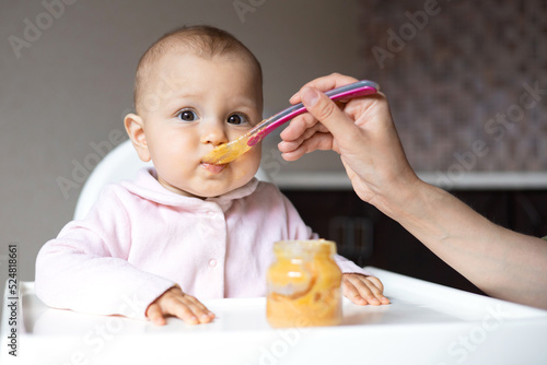 Baby food. A baby in a high chair eats vegetable puree from a spoon. Mom feeds the baby from a jar. Kitchen. Lifestyle.