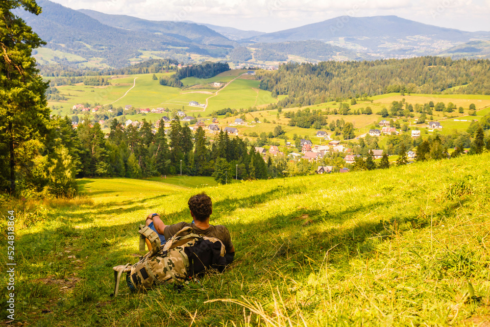 Młody mężczyzna z plecakiem siedzący na skraju lasu patrzący na piękny krajobraz w górach - obrazy, fototapety, plakaty 