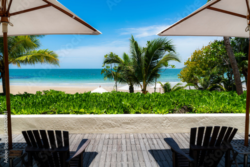A view of white sand beach and turquoise-color sea water from the perspective of wooden chair on the wooden deck sitting on the swimming pool side under the shade of white-big beach umbrella