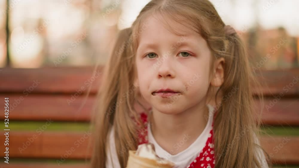 Preschooler girl with ponytails eats ice cream with pleasure sitting on wooden bench closeup. Kid bites cold dessert at sunset. Delicious sundae snack Preschooler girl with ponytails eats ice cream with pleasure sitting on wooden bench closeup. Kid bites cold dessert at sunset. Delicious sundae snack