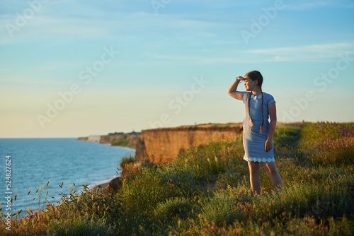 A school-age girl in a striped dress stands at sunset against the background of the sea.