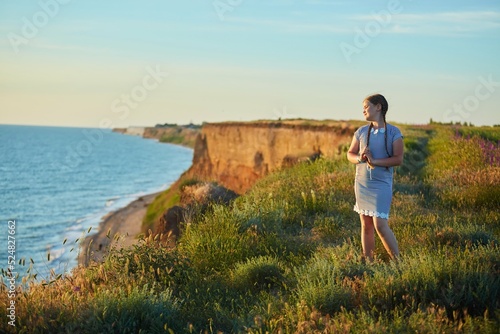 A school-age girl in a striped dress stands at sunset against the background of the sea. © Fotoproff