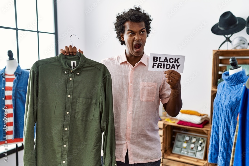 African man with curly hair holding black friday banner at retail shop angry and mad screaming frustrated and furious, shouting with anger. rage and aggressive concept. African man with curly hair holding black friday banner at retail shop angry and mad screaming frustrated and furious, shouting with anger. rage and aggressive concept.