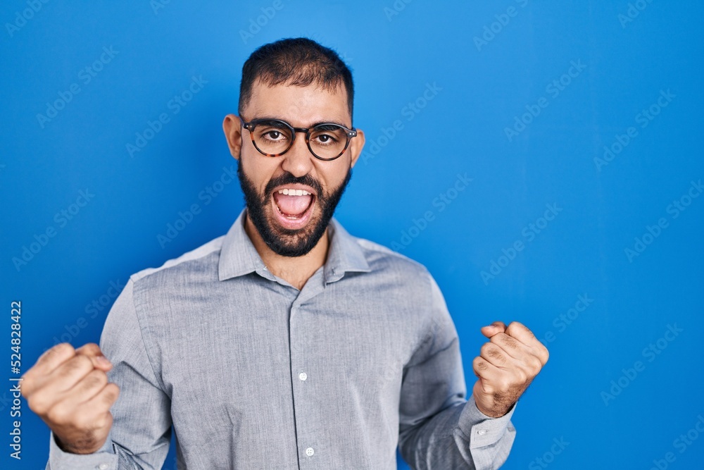 Middle east man with beard standing over blue background very happy and excited doing winner gesture with arms raised, smiling and screaming for success. celebration concept. Middle east man with beard standing over blue background very happy and excited doing winner gesture with arms raised, smiling and screaming for success. celebration concept.