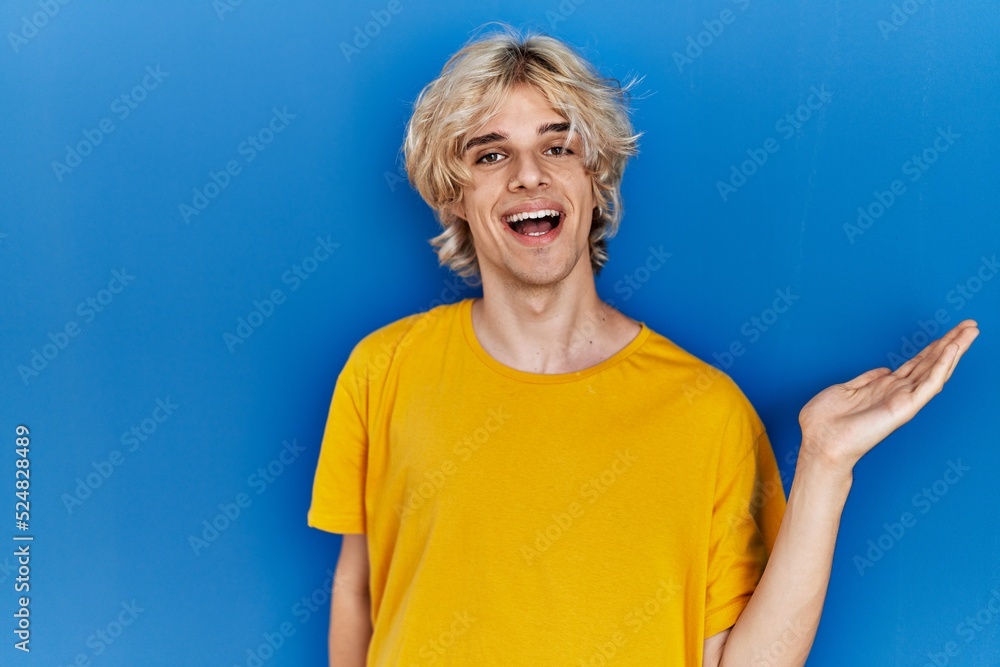 Young modern man standing over blue background smiling cheerful presenting and pointing with palm of hand looking at the camera. Young modern man standing over blue background smiling cheerful presenting and pointing with palm of hand looking at the camera.