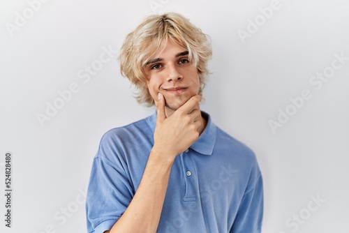 Young modern man standing over isolated background looking confident at the camera with smile with crossed arms and hand raised on chin. thinking positive.