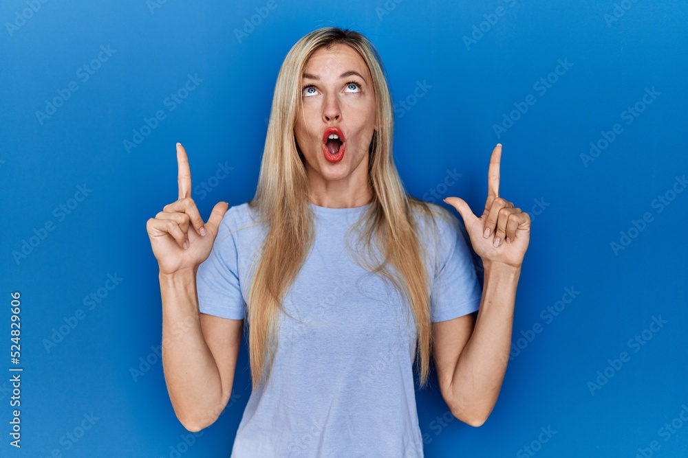 Beautiful blonde woman wearing casual t shirt over blue background amazed and surprised looking up and pointing with fingers and raised arms. Beautiful blonde woman wearing casual t shirt over blue background amazed and surprised looking up and pointing with fingers and raised arms.