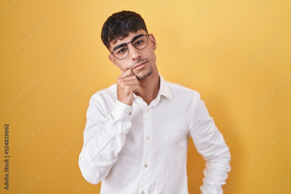 Fototapeta premium Young hispanic man standing over yellow background with hand on chin thinking about question, pensive expression. smiling with thoughtful face. doubt concept.