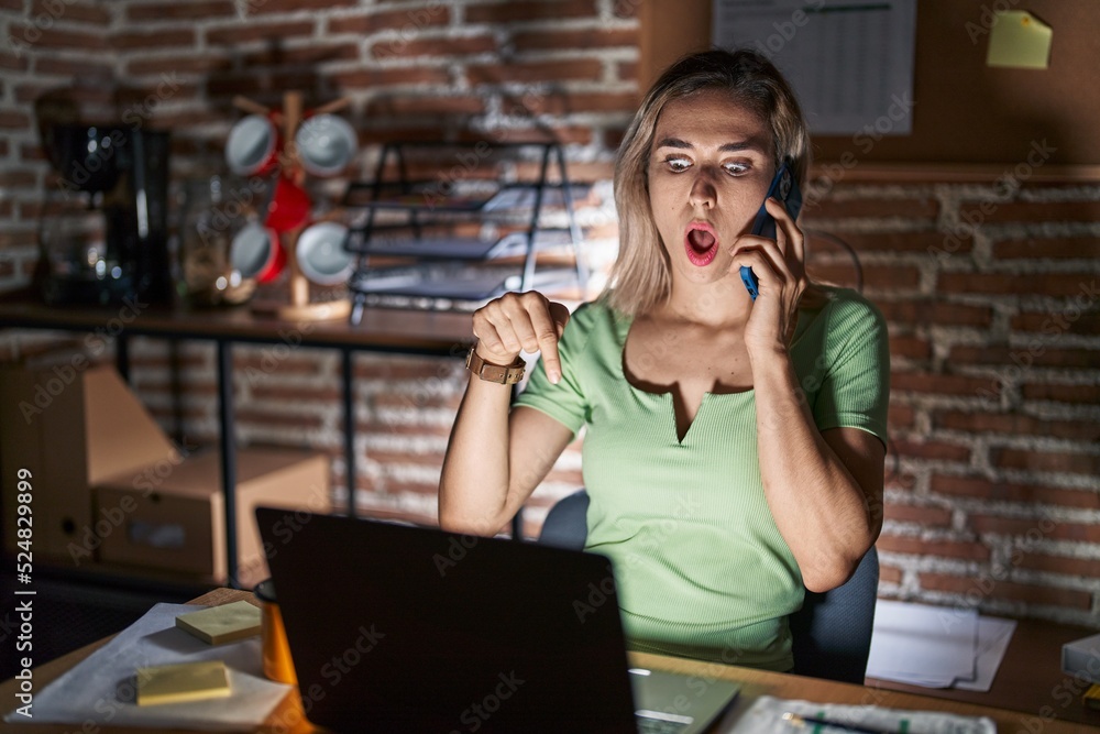 Young beautiful woman working at the office at night speaking on the phone pointing down with fingers showing advertisement, surprised face and open mouth Young beautiful woman working at the office at night speaking on the phone pointing down with fingers showing advertisement, surprised face and open mouth
