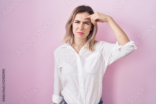Young beautiful woman standing over pink background pointing unhappy to pimple on forehead, ugly infection of blackhead. acne and skin problem