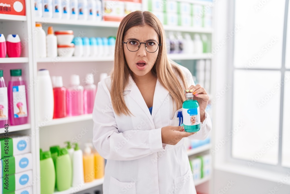 Young blonde woman working at pharmacy drugstore holding syrup afraid and shocked with surprise and amazed expression, fear and excited face. Young blonde woman working at pharmacy drugstore holding syrup afraid and shocked with surprise and amazed expression, fear and excited face.