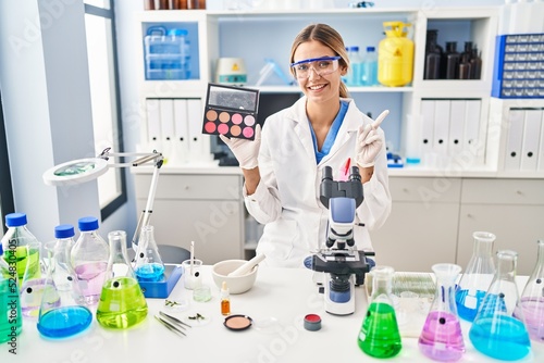 Young blonde woman working at scientist laboratory with make up smiling happy pointing with hand and finger to the side