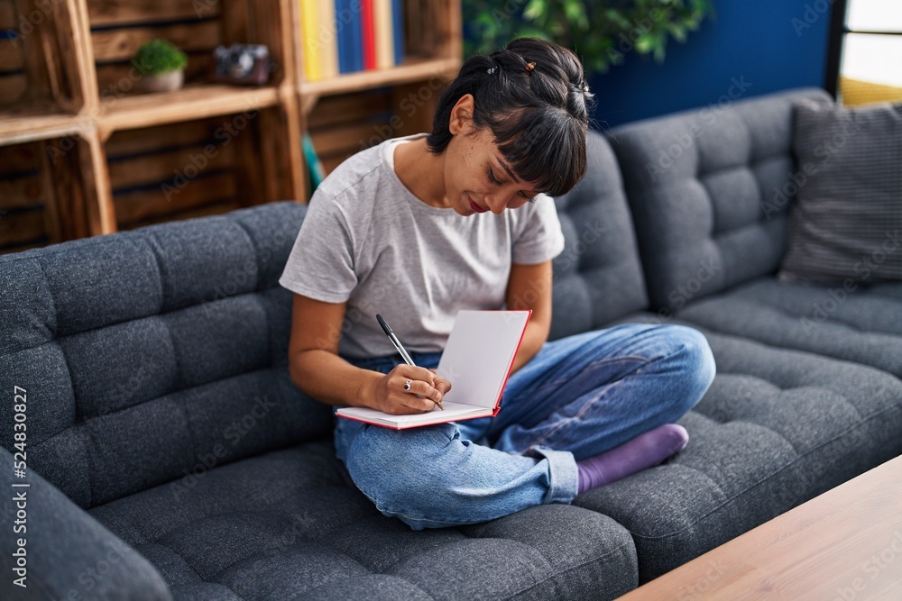 Young woman writing on book sitting on sofa at home