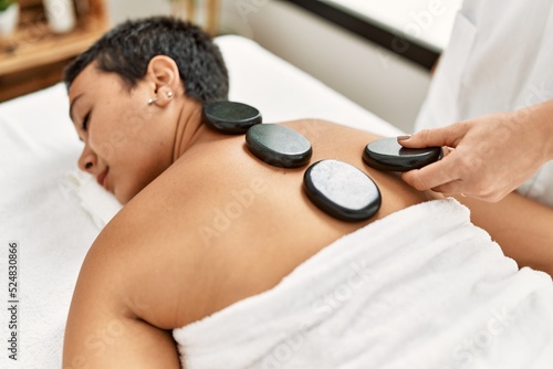 Young hispanic woman having back massage using black hot stones at beauty center