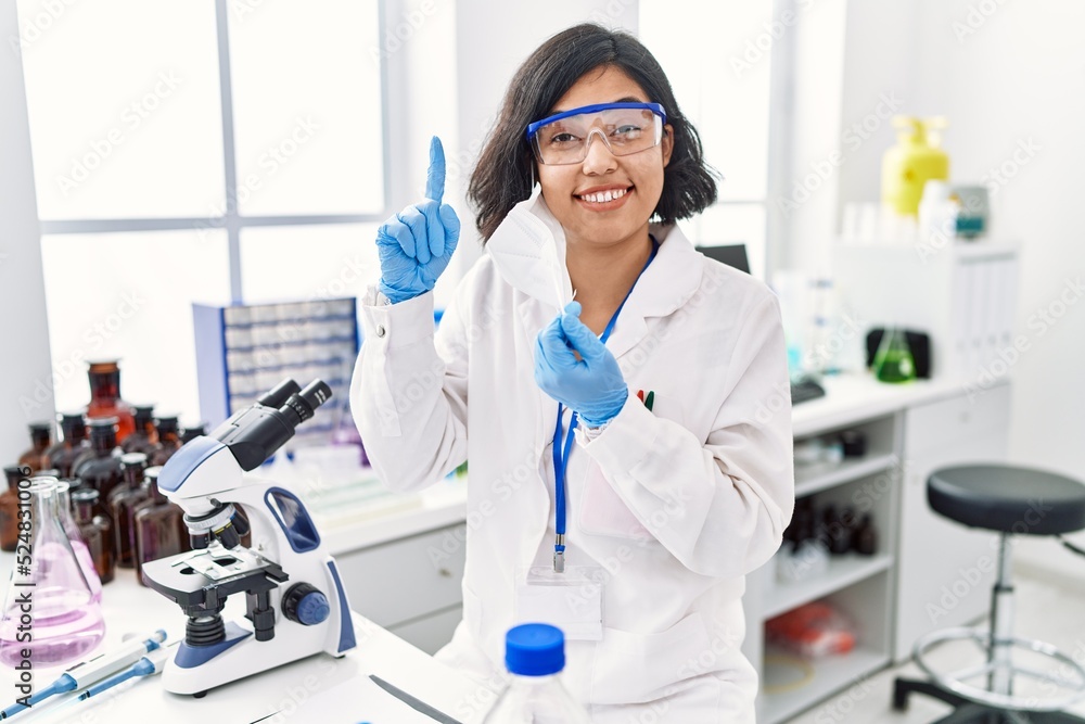 Young hispanic woman working at scientist laboratory with safety mask smiling with an idea or question pointing finger with happy face, number one Young hispanic woman working at scientist laboratory with safety mask smiling with an idea or question pointing finger with happy face, number one
