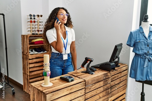 Young latin shopkeeper woman talking on the smartphone working at clothing store.