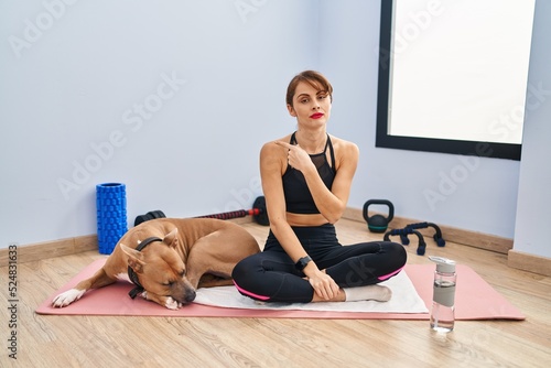 Young beautiful woman sitting on yoga mat pointing with hand finger to the side showing advertisement, serious and calm face