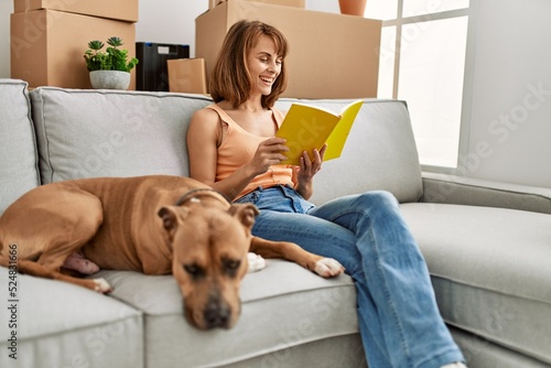 Young caucasian woman reading book sitting on sofa with dog at home