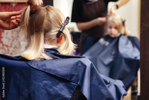 Hair salon, barber woman make fashionable pretty hairstyle for cute young blond child in modern barbershop. Hairdresser makes hairdo for little girl in barber shop. Concept hairstyle and beauty