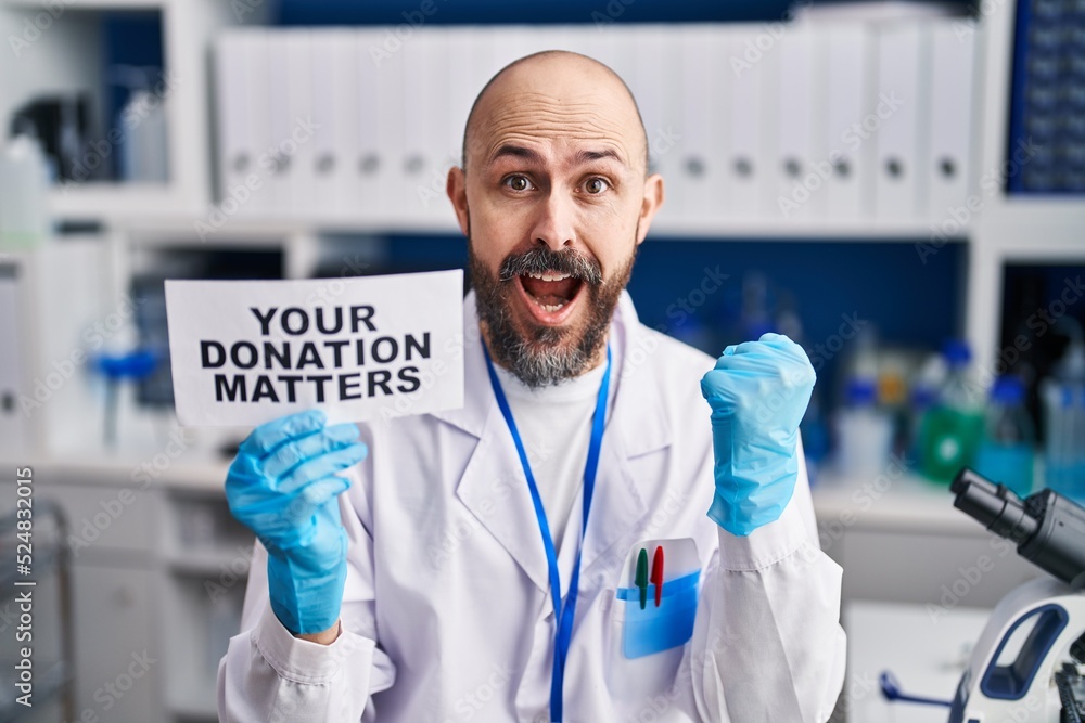 Young hispanic man working at scientist laboratory holding you donation matters banner screaming proud, celebrating victory and success very excited with raised arms Young hispanic man working at scientist laboratory holding you donation matters banner screaming proud, celebrating victory and success very excited with raised arms