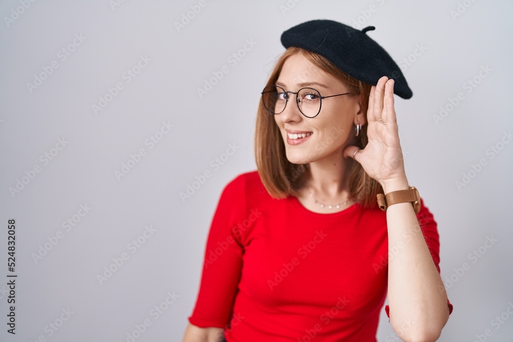 Young redhead woman standing wearing glasses and beret smiling with hand over ear listening an hearing to rumor or gossip. deafness concept. Young redhead woman standing wearing glasses and beret smiling with hand over ear listening an hearing to rumor or gossip. deafness concept.
