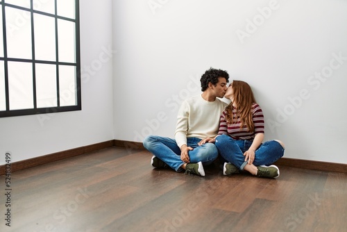 Young hispanic couple kissing and sitting on the floor at empty new home.