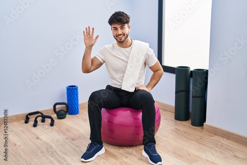 Hispanic man with beard sitting on pilate balls at yoga room showing and pointing up with fingers number five while smiling confident and happy.