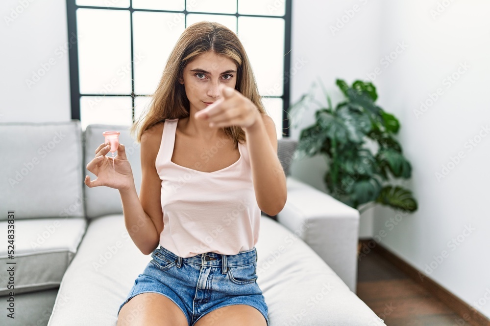 Young blonde woman holding menstrual cup pointing with finger to the camera and to you, confident gesture looking serious Young blonde woman holding menstrual cup pointing with finger to the camera and to you, confident gesture looking serious