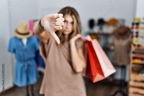 Young blonde woman holding shopping bags at retail shop looking unhappy and angry showing rejection and negative with thumbs down gesture. bad expression.