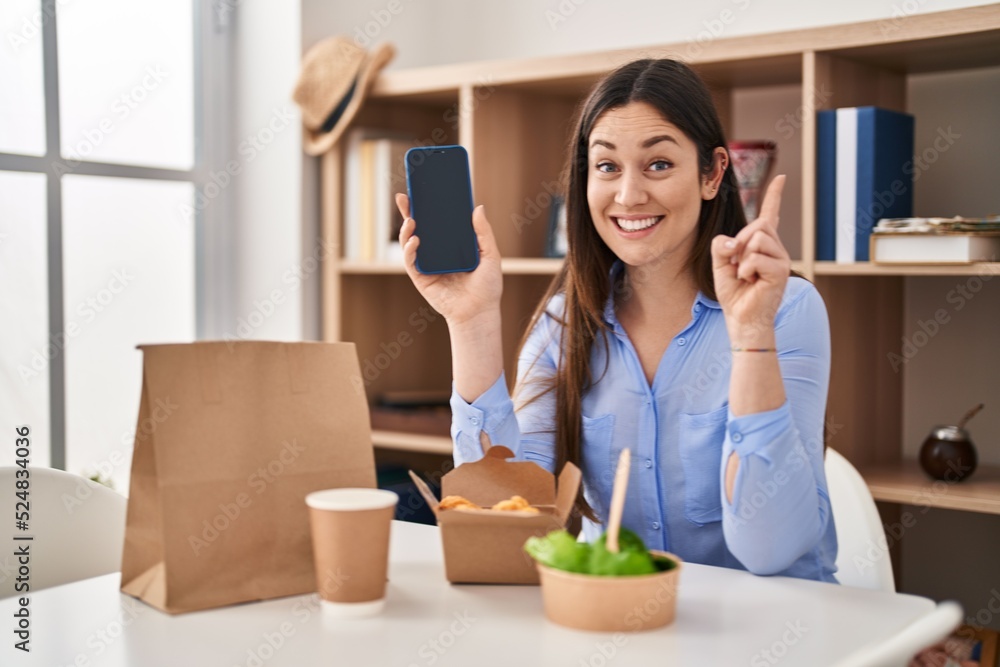 Young brunette woman eating take away food at home showing smartphone screen surprised with an idea or question pointing finger with happy face, number one Young brunette woman eating take away food at home showing smartphone screen surprised with an idea or question pointing finger with happy face, number one