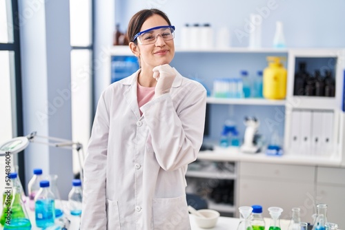 Young brunette woman working at scientist laboratory smiling looking confident at the camera with crossed arms and hand on chin. thinking positive.