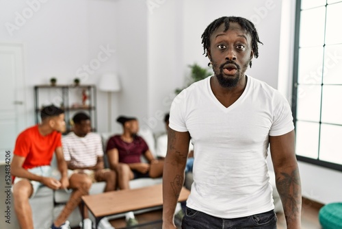Young african man with friends at the living room scared and amazed with open mouth for surprise  disbelief face