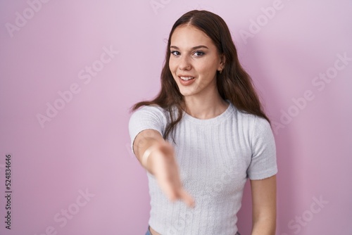 Young hispanic girl standing over pink background smiling friendly offering handshake as greeting and welcoming. successful business.
