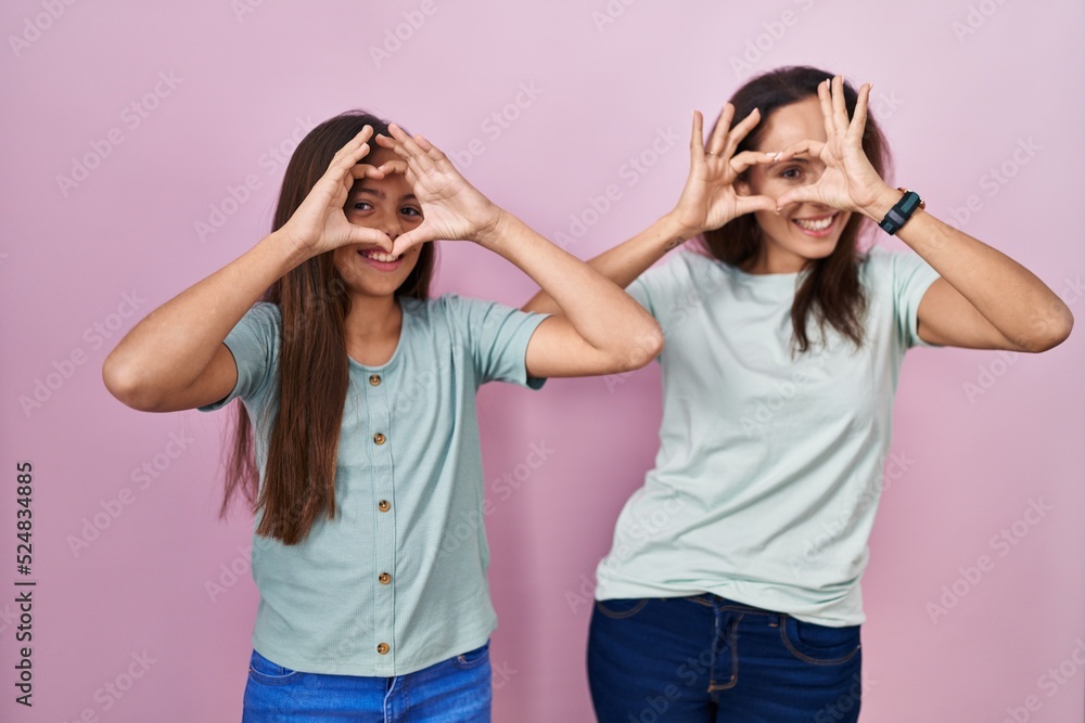 Young mother and daughter standing over pink background doing heart shape with hand and fingers smiling looking through sign Young mother and daughter standing over pink background doing heart shape with hand and fingers smiling looking through sign