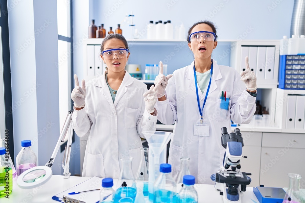 Young mother and daughter at scientist laboratory amazed and surprised looking up and pointing with fingers and raised arms. Young mother and daughter at scientist laboratory amazed and surprised looking up and pointing with fingers and raised arms.