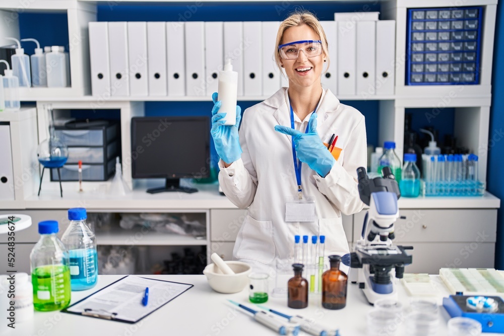 Young caucasian woman working at scientist laboratory holding body lotion smiling happy pointing with hand and finger