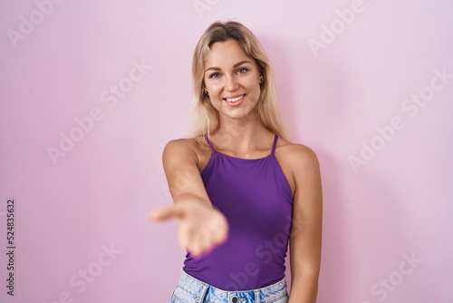 Young blonde woman standing over pink background smiling friendly offering handshake as greeting and welcoming. successful business.