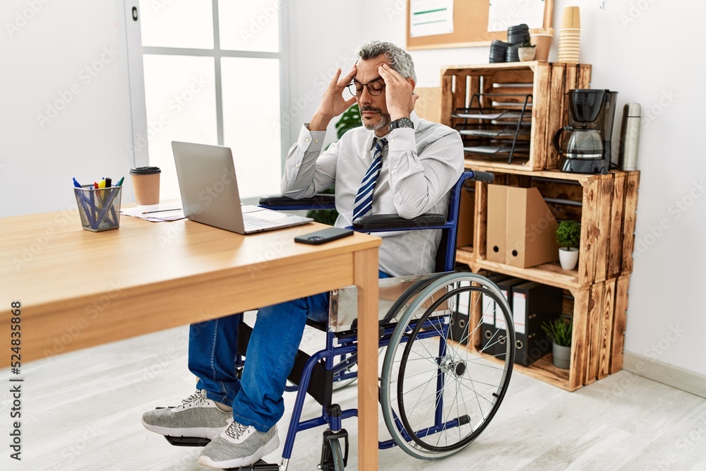 Middle age hispanic man working at the office sitting on wheelchair with hand on head for pain in head because stress. suffering migraine. Middle age hispanic man working at the office sitting on wheelchair with hand on head for pain in head because stress. suffering migraine.