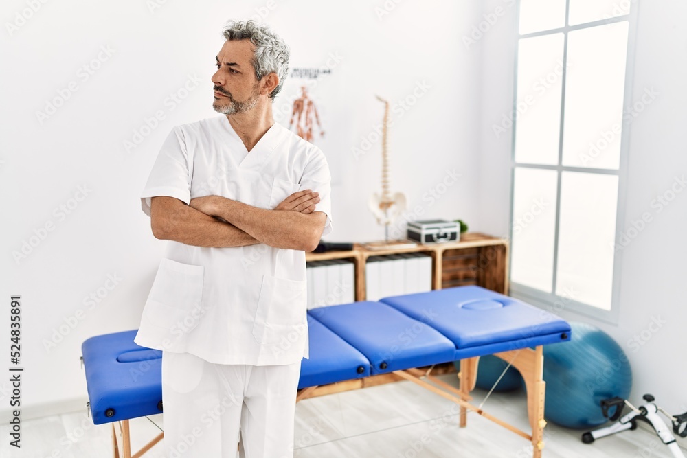 Middle age hispanic therapist man working at pain recovery clinic looking to the side with arms crossed convinced and confident Middle age hispanic therapist man working at pain recovery clinic looking to the side with arms crossed convinced and confident
