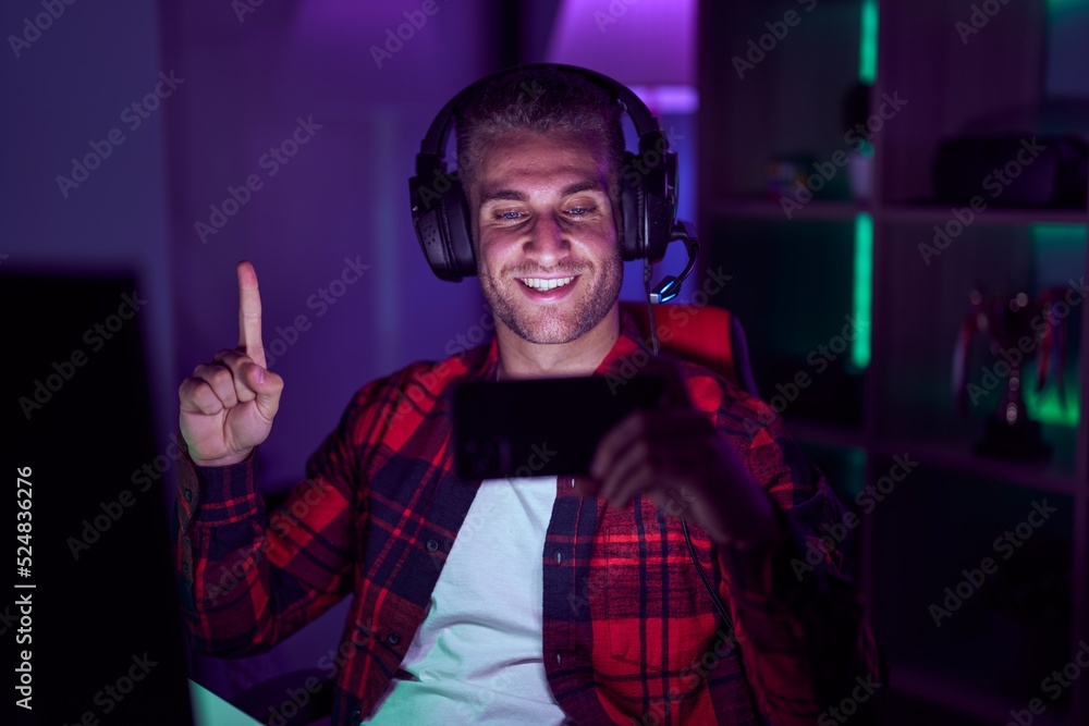 Young caucasian man playing video games with smartphone smiling with an idea or question pointing finger with happy face, number one Young caucasian man playing video games with smartphone smiling with an idea or question pointing finger with happy face, number one