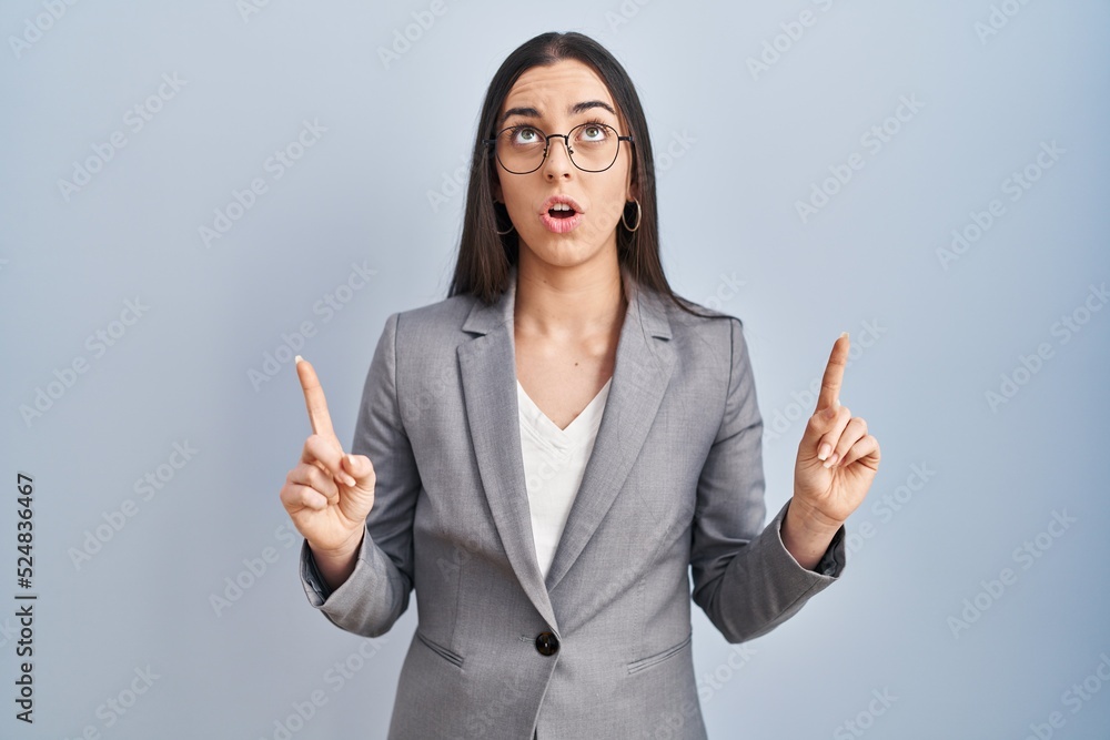 Hispanic business woman wearing glasses amazed and surprised looking up and pointing with fingers and raised arms. Hispanic business woman wearing glasses amazed and surprised looking up and pointing with fingers and raised arms.