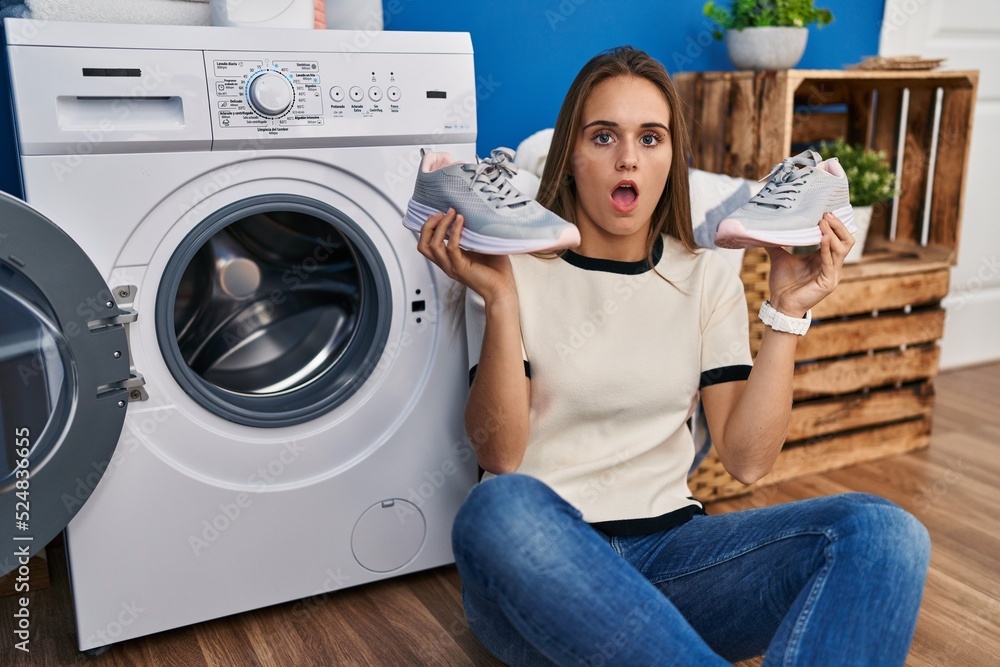 Young woman putting sneakers in washing machine in shock face, looking skeptical and sarcastic, surprised with open mouth Young woman putting sneakers in washing machine in shock face, looking skeptical and sarcastic, surprised with open mouth