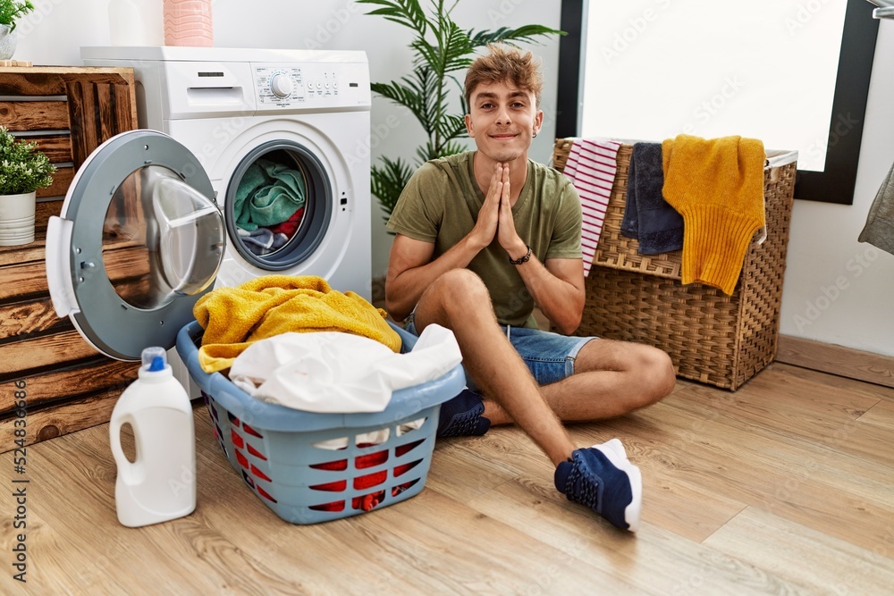 Young caucasian man putting dirty laundry into washing machine praying with hands together asking for forgiveness smiling confident. Young caucasian man putting dirty laundry into washing machine praying with hands together asking for forgiveness smiling confident.