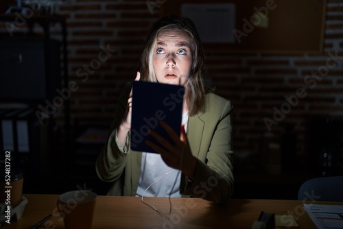 Blonde caucasian woman working at the office at night pointing up looking sad and upset, indicating direction with fingers, unhappy and depressed.