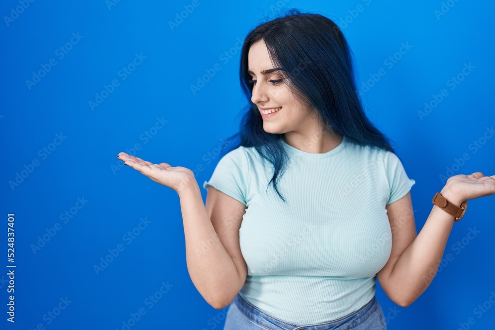 Young modern girl with blue hair standing over blue background smiling showing both hands open palms, presenting and advertising comparison and balance Young modern girl with blue hair standing over blue background smiling showing both hands open palms, presenting and advertising comparison and balance