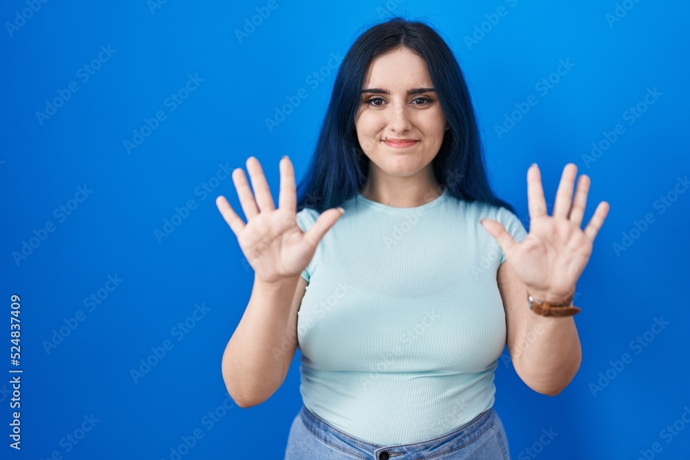 Young modern girl with blue hair standing over blue background showing and pointing up with fingers number ten while smiling confident and happy. Young modern girl with blue hair standing over blue background showing and pointing up with fingers number ten while smiling confident and happy.