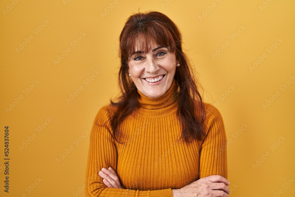 Middle age hispanic woman standing over yellow background happy face smiling with crossed arms looking at the camera. positive person. Middle age hispanic woman standing over yellow background happy face smiling with crossed arms looking at the camera. positive person.